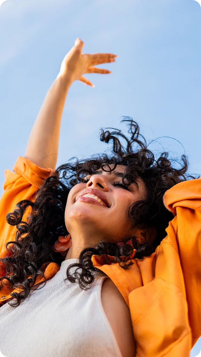 Joyful woman with curly hair in orange jacket reaching toward sky against blue background - in Carmel, IN