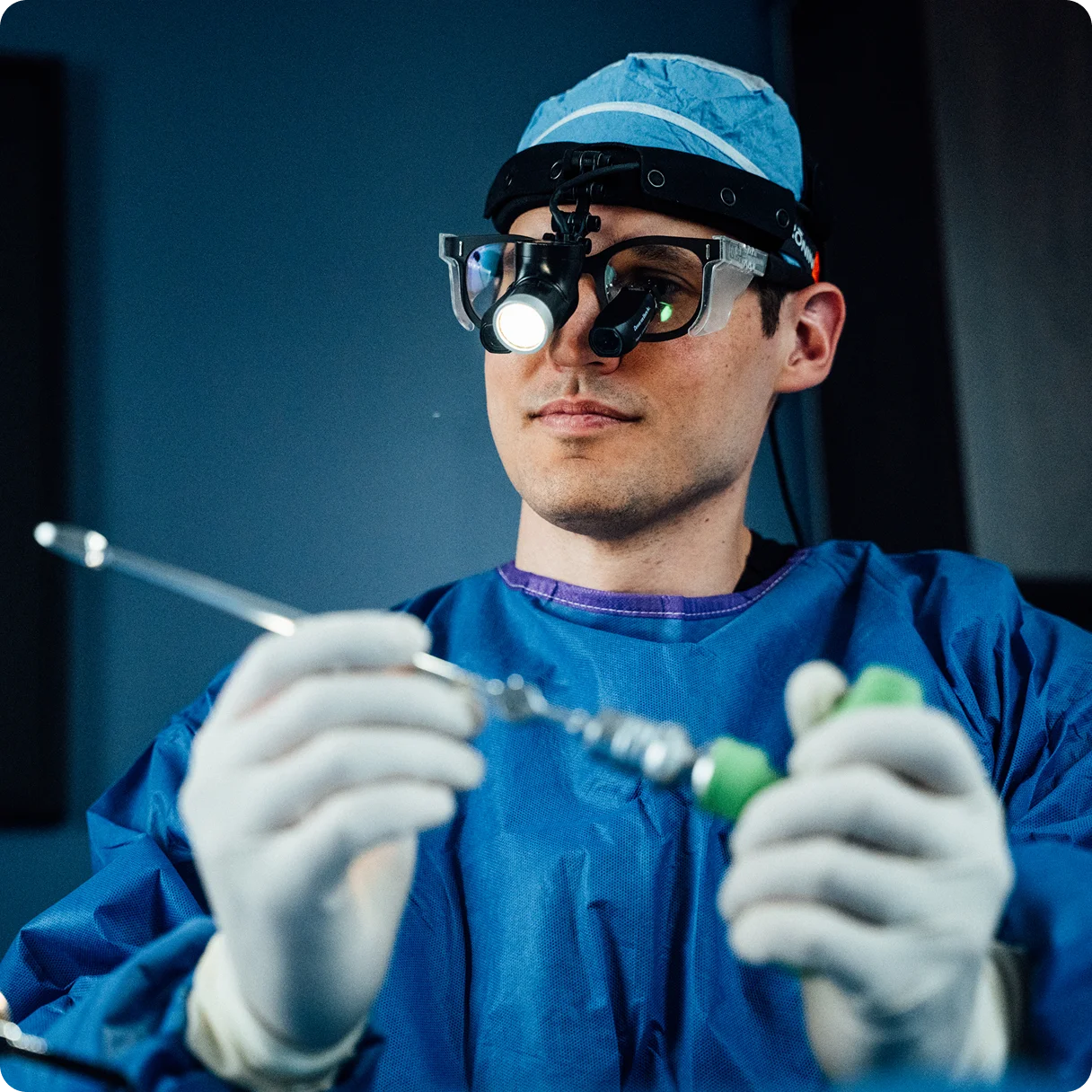 Dr Peter Swiatek MD in blue uniform and magnifying glasses inspecting medical device in operating room setting - in Carmel, IN