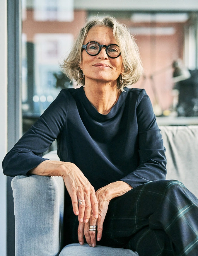 Elegant mature woman with gray hair and glasses wearing black outfit sitting on modern sofa in bright contemporary office - ALIF in Carmel, IN