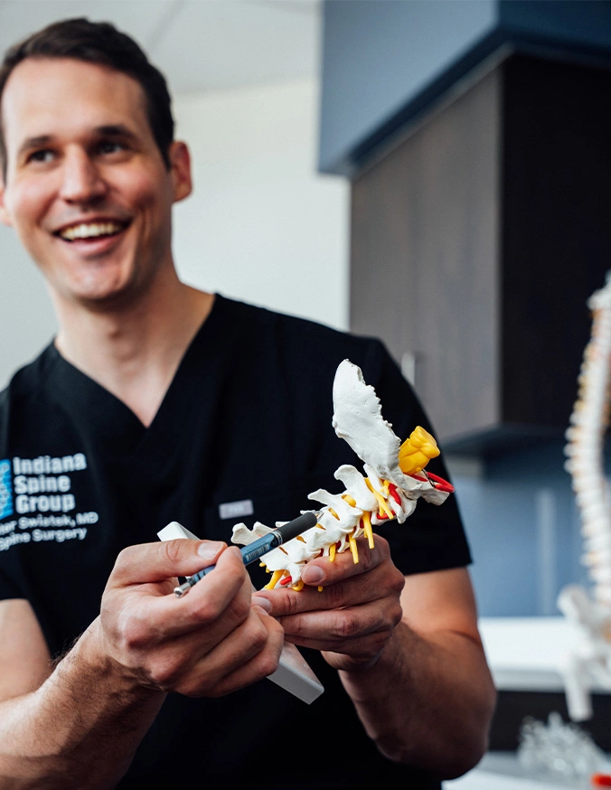Dr Peter Swiatek MD smiling while holding spine model in hands, wearing black scrubs - Cervical disc herniation in Carmel, IN