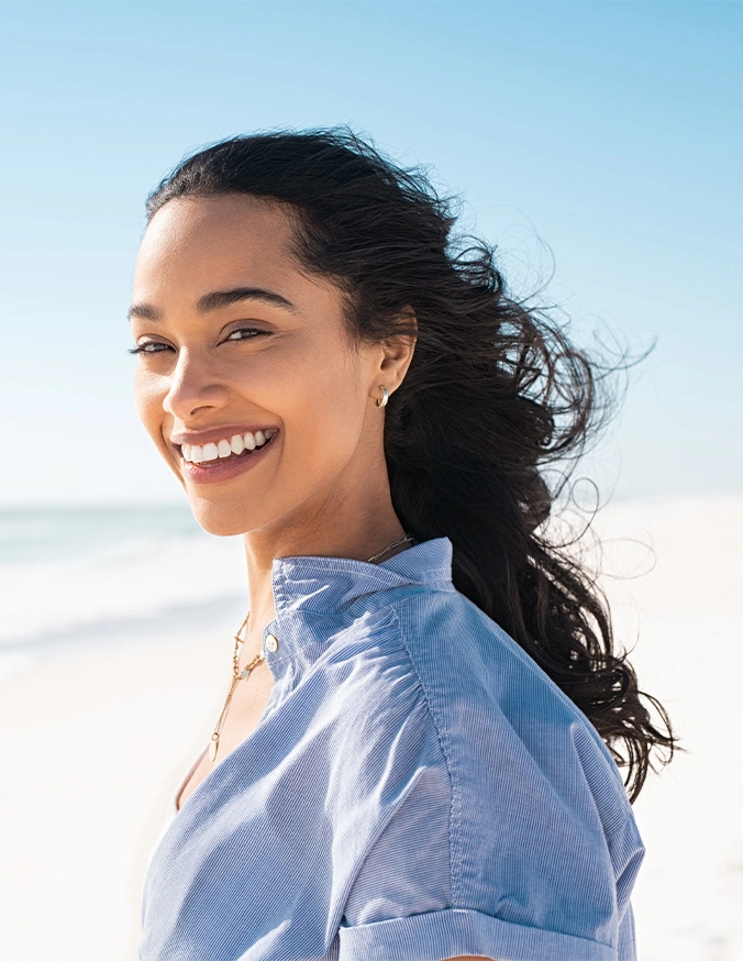 Smiling woman with long dark hair on beach wearing blue shirt in natural sunlight - Cervical spondylolisthesis in Carmel, IN