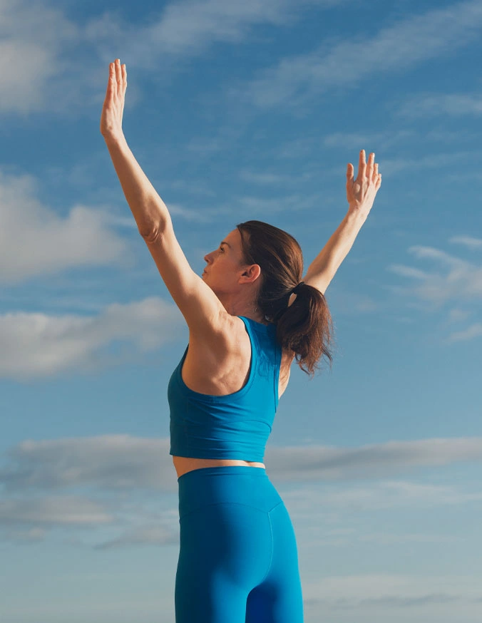 Woman in blue athletic wear stretching arms overhead against sky, demonstrating back flexibility and spinal health exercise - Lumbar degenerative disc disease in Carmel, IN