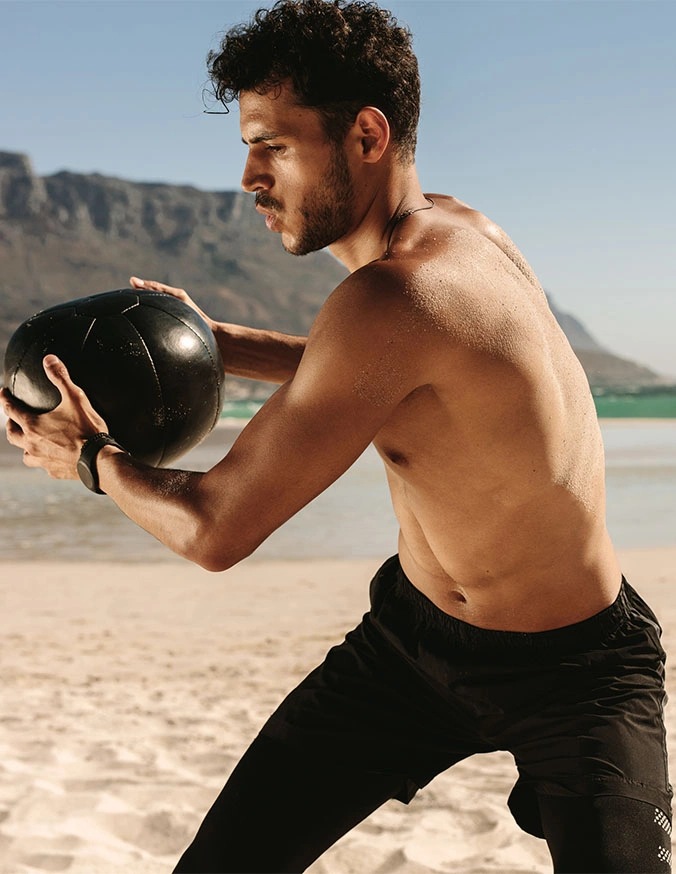Shirtless athletic man training with medicine ball on beach with mountain backdrop - Lumbar disc heritations in Carmel, IN