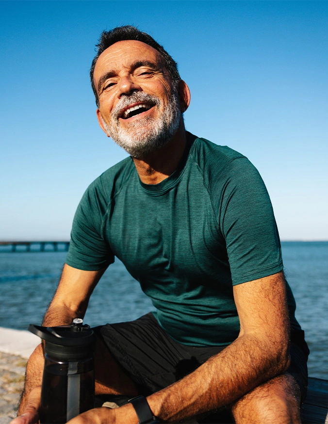 Smiling mature man in teal shirt sitting by waterfront on sunny day with bridge in background - Lumbar Laminectomy in Carmel, IN