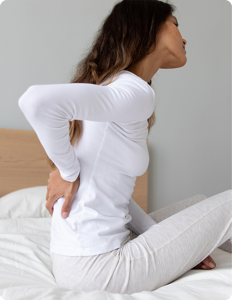 Woman experiencing lower back pain while sitting on bed, holding her lumbar spine area - Tilf in Carmel, IN